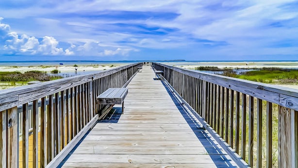 Ein Holzsteg führt zu einem Strand, der unter blauem Himmel liegt.
