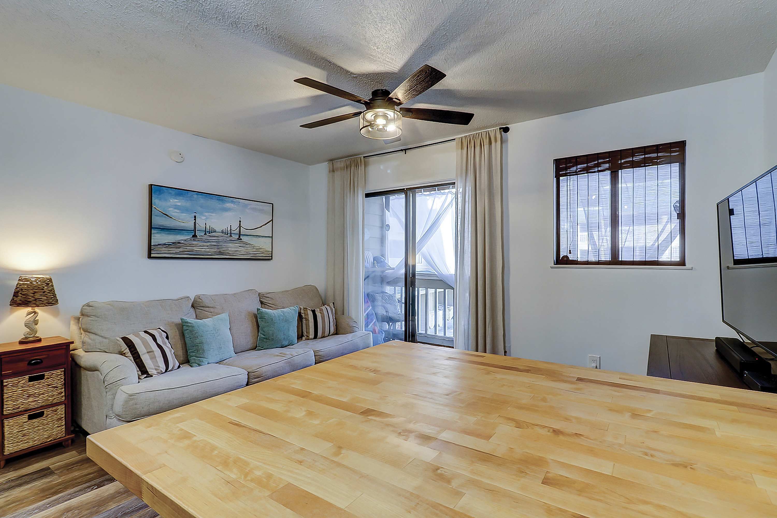 A room with a ceiling fan, a large wooden table, a sofa with cushions, a TV, and a window with curtains.