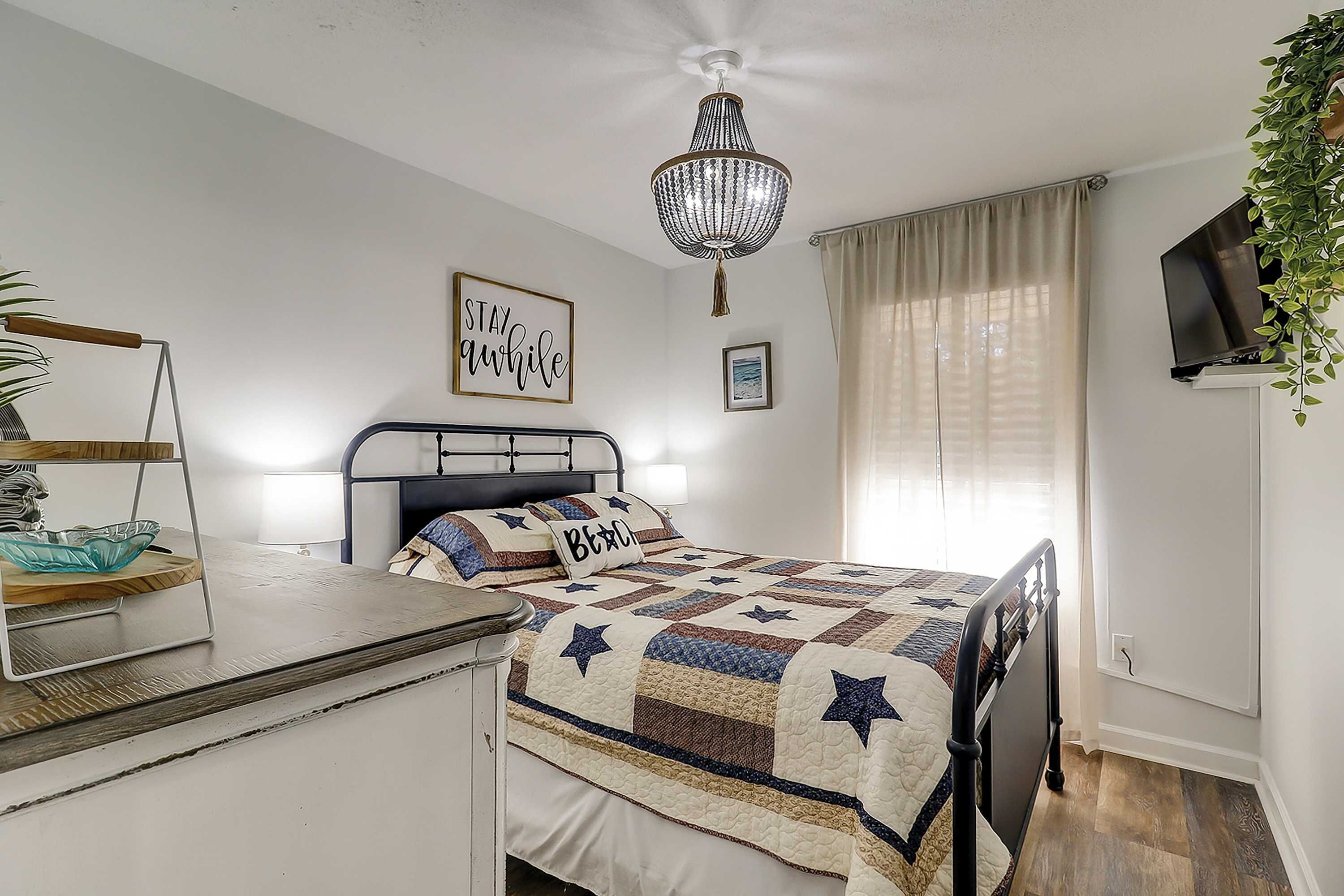 Un dormitorio con cama, mesita de noche, planta colgante, candelabro, televisor y ventana con cortinas.