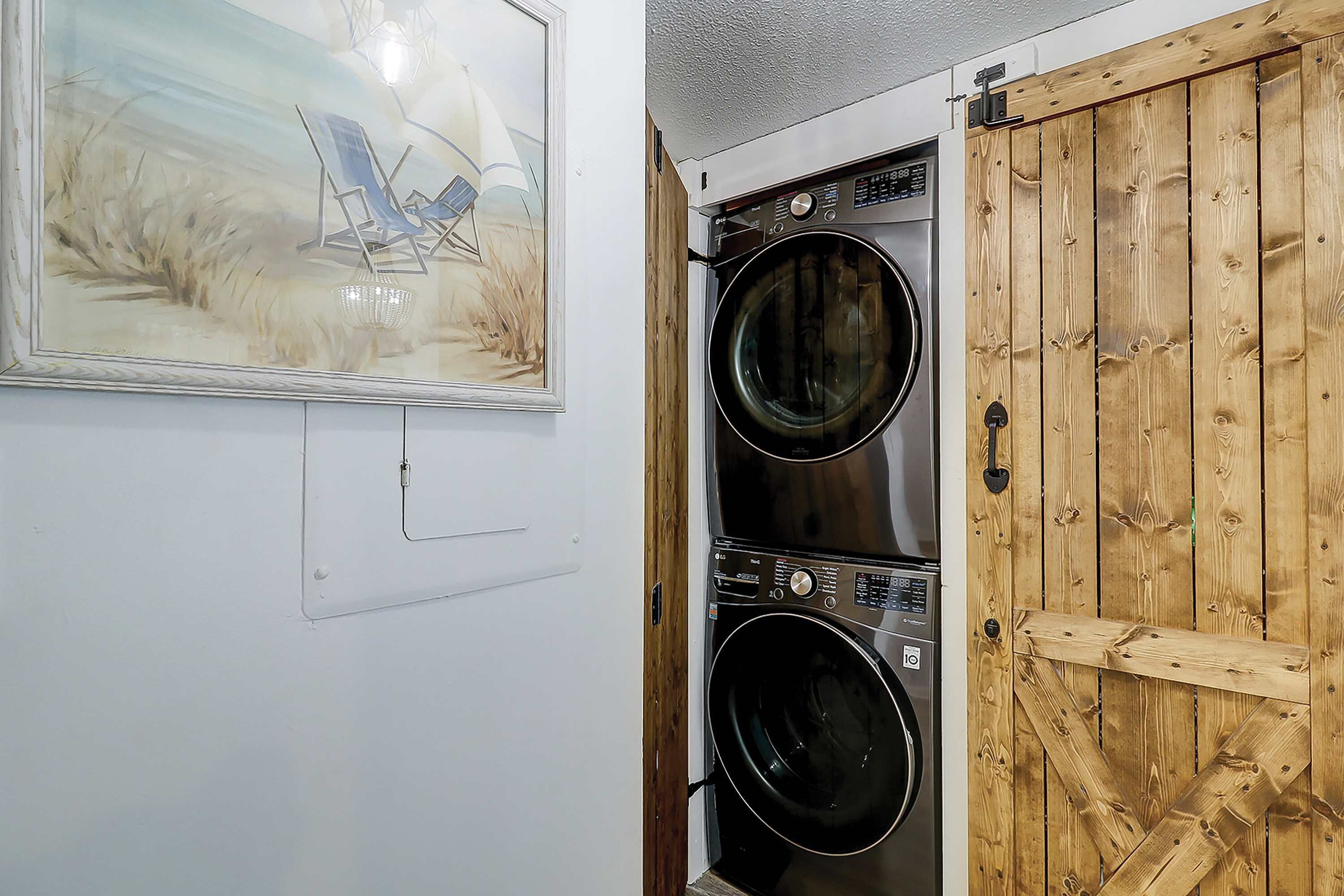 A laundry room with two stacked front-loading washers and dryers, a wooden door, and a beach-themed painting on the wall.