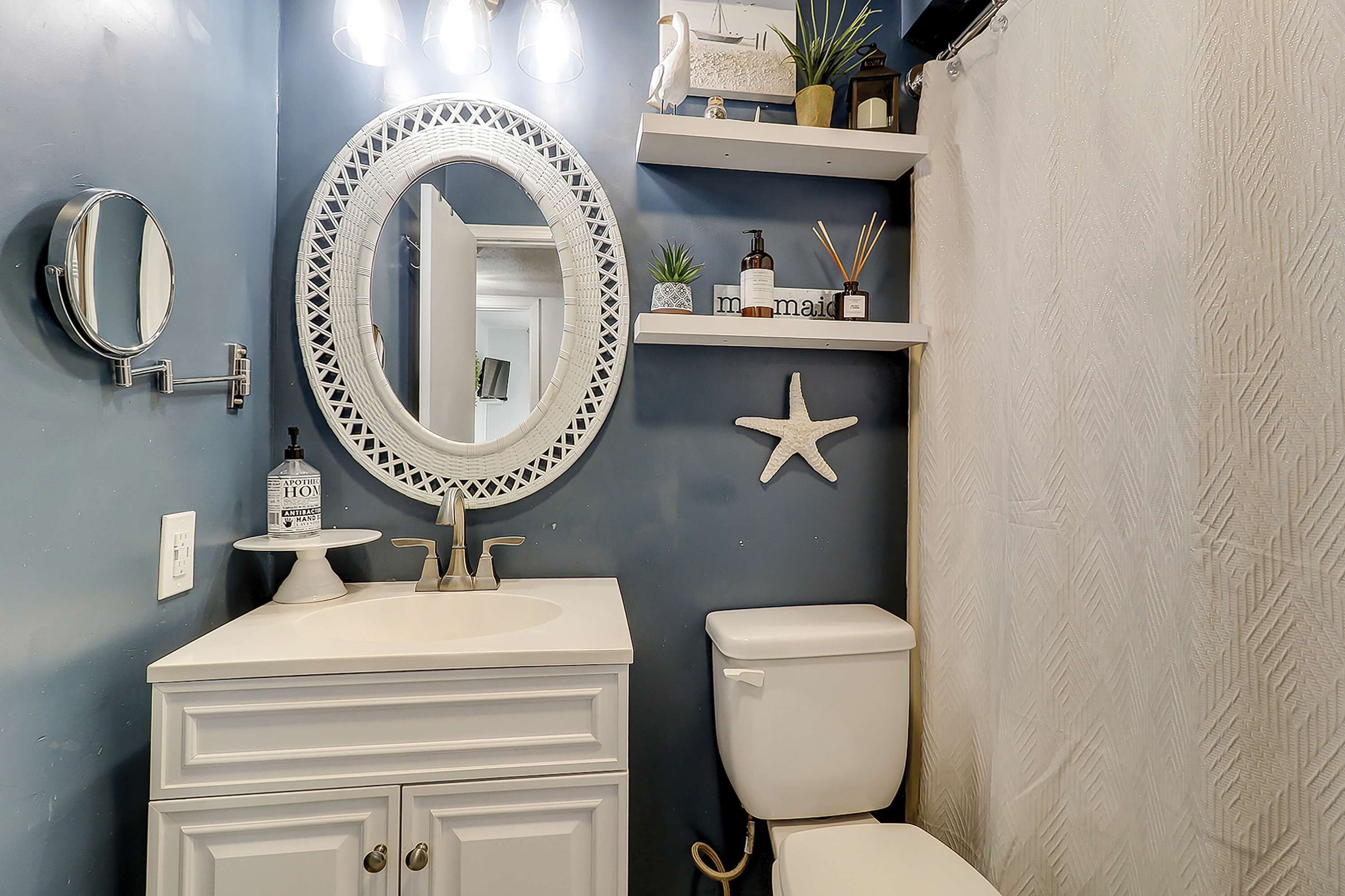 A bathroom with a white vanity, a round mirror, a toilet, and a shelf with toiletries.