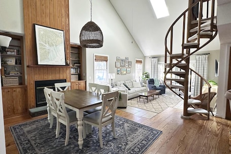 A spacious living area with a dining table, white chairs, a fireplace, a bookshelf, a staircase, and a large window.
