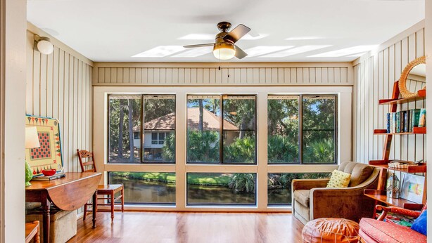 A spacious room with large windows, a ceiling fan, a wooden dining table, and a cozy seating area. - Vibrant, Waterfront Home with Free WiFi, Full Kitchen, Gas Grill, & A/C (Edisto Beach)