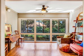 A spacious room with large windows, a ceiling fan, a wooden dining table, and a cozy seating area.