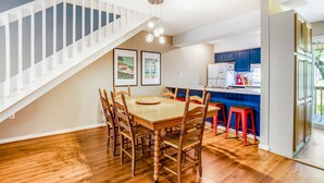 A dining area with a wooden table and chairs, a kitchen with blue cabinets, and a staircase. - Vibrant, Waterfront Home with Free WiFi, Full Kitchen, Gas Grill, & A/C (Edisto Beach)