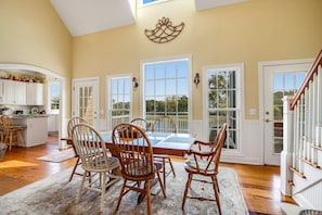A dining area with a table and chairs, large windows, and a kitchen in the background. - Charming Home w/ Lagoon View, WiFi, Laundry, Central AC - Walk to the Beach (Edisto Beach)