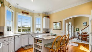 A kitchen with white cabinetry, a central island with bar stools, and a view through large windows. - Charming Home w/ Lagoon View, WiFi, Laundry, Central AC - Walk to the Beach (Edisto Beach)