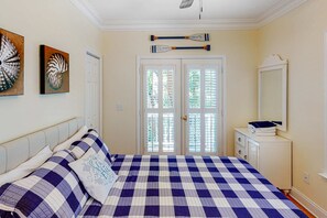 A bedroom with a checkered bedspread, two paintings on the wall, a mirror, and a door with shutters. - Sun-kissed home with AC, fireplace & W/D - short drive to the beach (Edisto Beach)