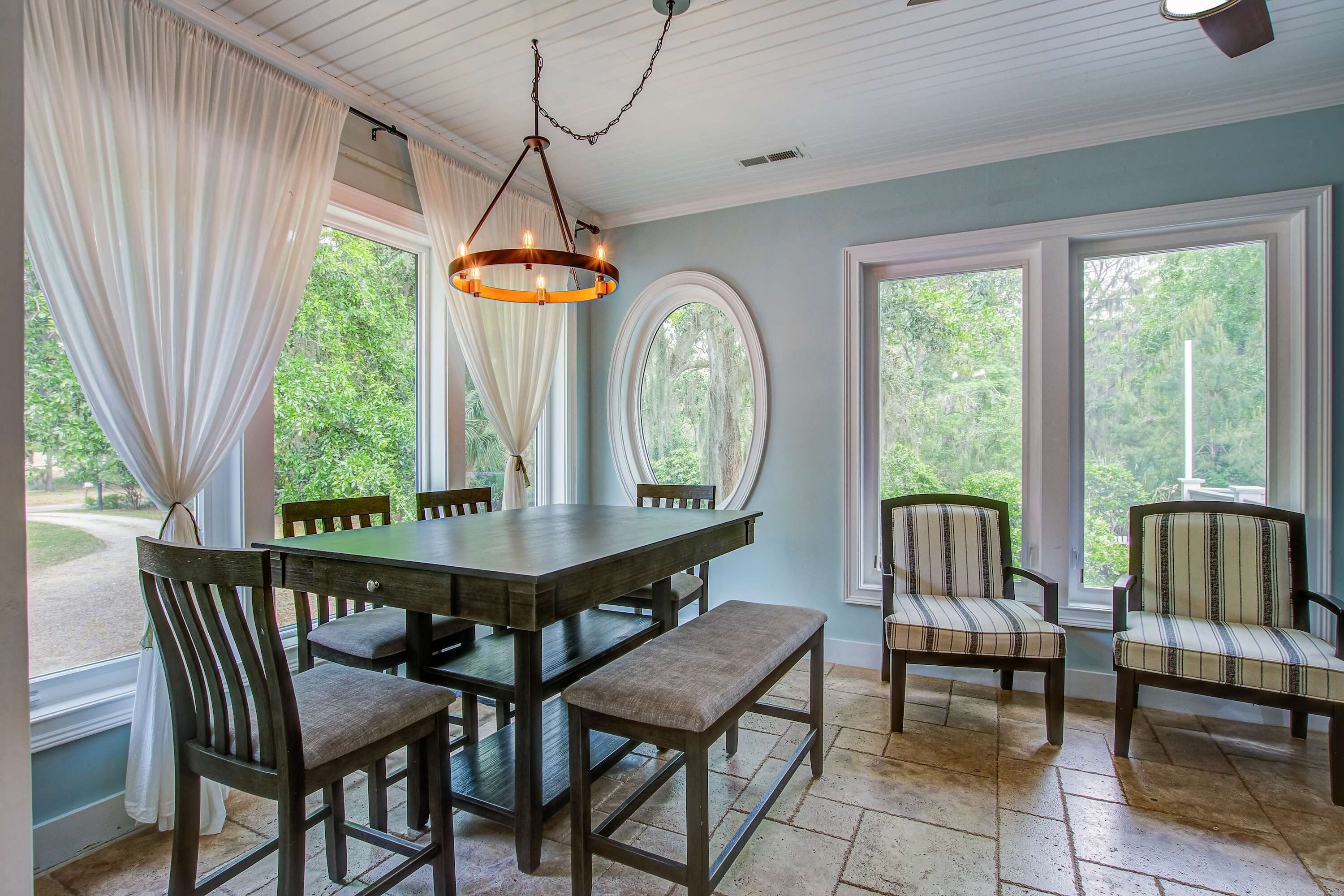 A dining area with a wooden table, chairs, and a bench. There are large windows with curtains, a chandelier, and a view of greenery outside.