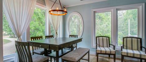 A dining area with a wooden table, chairs, and a bench. There are large windows with curtains, a chandelier, and a view of greenery outside.