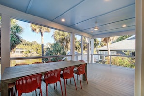 A covered outdoor dining area with a wooden table and red chairs, large windows, and a view of palm trees and buildings. - Modern house near oceanfront, EV charger & large screened-in porch (Edisto Beach)