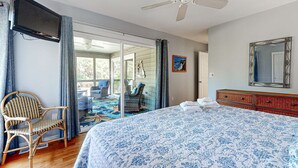Ein Schlafzimmer mit einem Bett, einem Sessel, einer Kommode, einem Deckenventilator und einer Glasschiebetür, die auf einen Balkon führt.