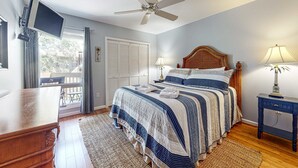 Ein Schlafzimmer mit Bett, Deckenventilator, Wand-TV, Nachttisch mit Lampe und Blick ins Freie durch ein Fenster.