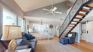Ein modernes Wohnzimmer mit einer Couch, einem Couchtisch, einer Lampe, einem Essbereich mit Stühlen, einer Küche im Hintergrund und einer Treppe mit Geländer.