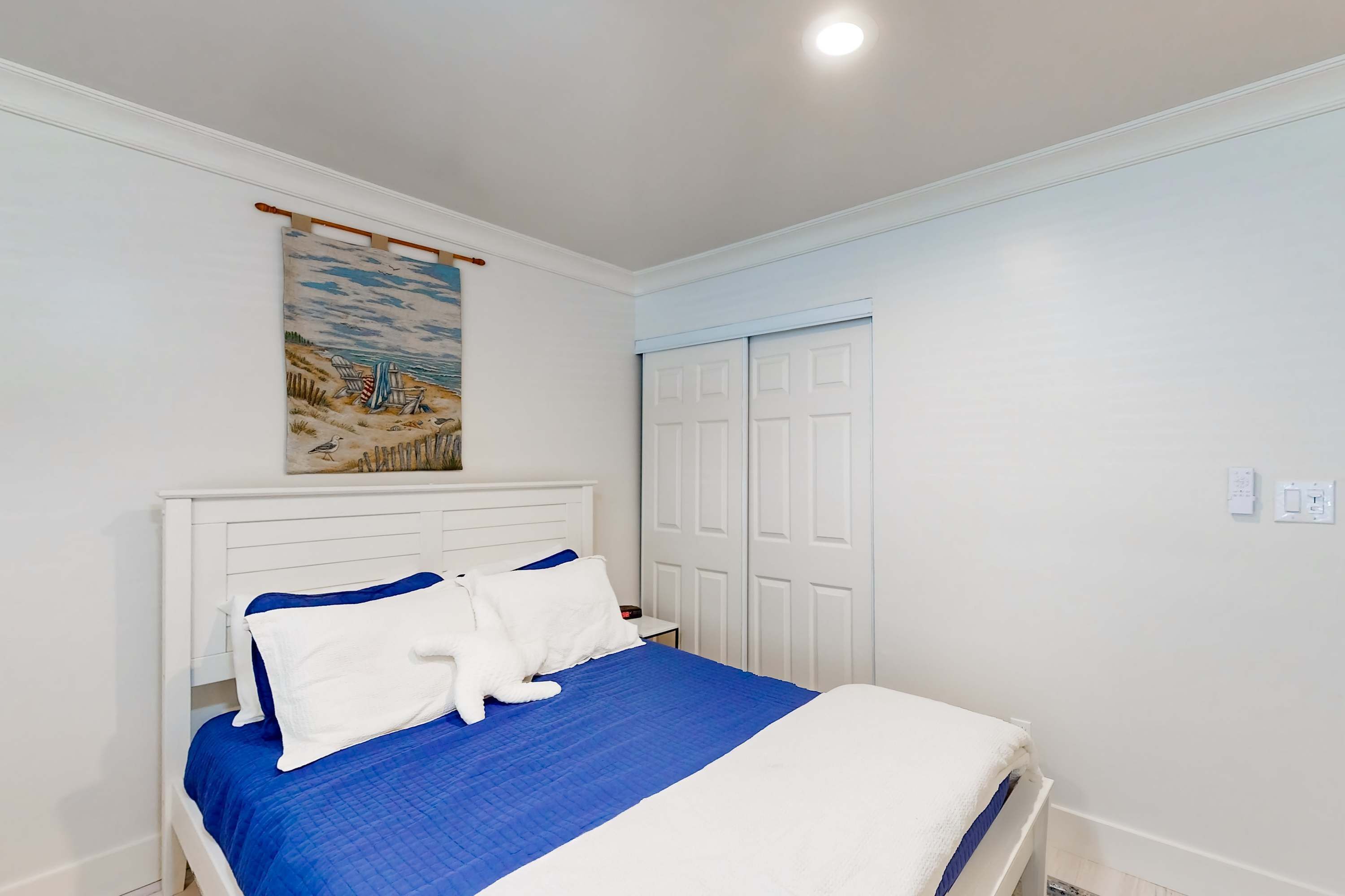 A neatly made bed with white and blue bedding, a white headboard, and a beach-themed painting above it.