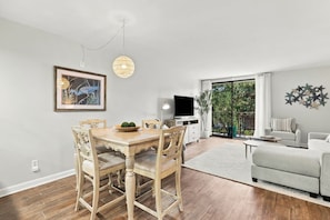 Een eetgedeelte met een houten tafel en stoelen, een schilderij, een kroonluchter en een televisie in een woonkamer met een balkon.