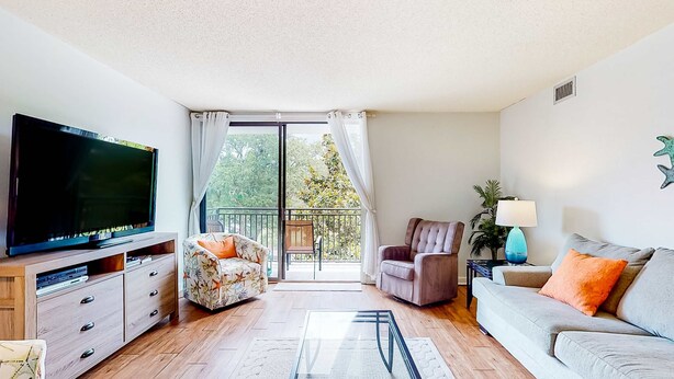 A living room with a flat-screen TV, a sofa, a chair, a small table with a lamp, and a view of a balcony.