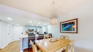 A dining area with a wooden table and chairs, a hanging light fixture, and a kitchen in the background.