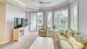 Un salon doté d’un téléviseur à écran plat, d’un ventilateur de plafond, d’un fauteuil en osier, d’une table basse en verre et d’un canapé agrémenté de coussins aux motifs tropicaux.