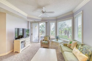 Ein Wohnzimmer mit Flachbildfernseher, Deckenventilator, Korbstuhl, Glastisch und einer Couch mit tropischen Kissen.