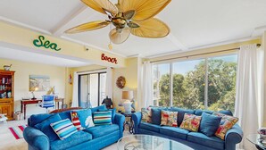 Ein Wohnzimmer mit zwei blauen Sofas, einem Glastisch, einem Deckenventilator und einem großen Fenster mit Blick auf Bäume.