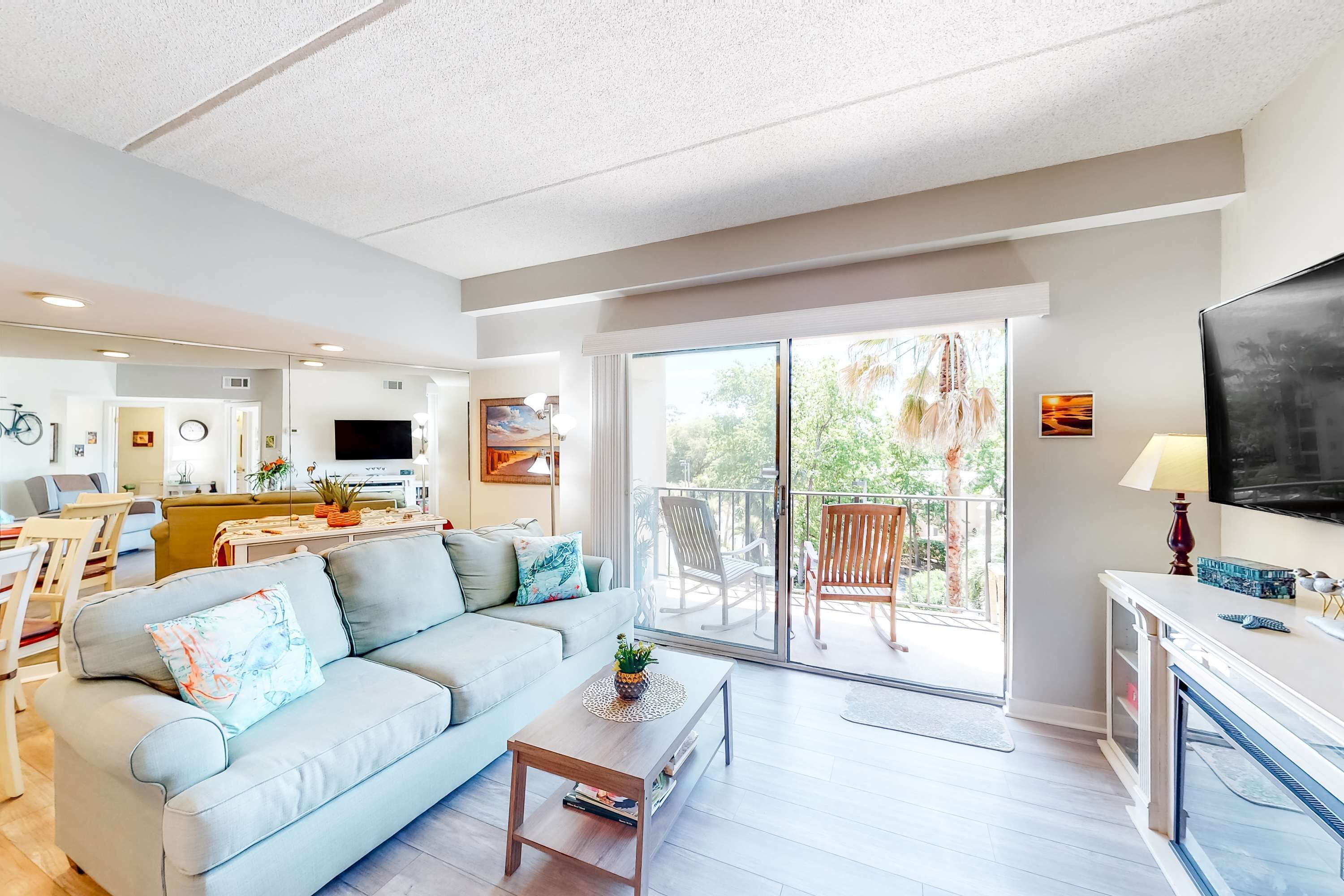 A modern living room with a sofa, coffee table, and a TV. There is a balcony with chairs and a dining area in the background.