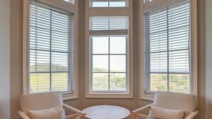 Una habitación con una mesa redonda y dos sillas, ventanas amplias con persianas y vistas a la vegetación exterior.