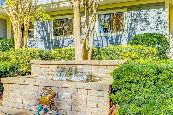 A landscaped garden with a water feature, a crab sculpture, and a building with windows in the background. - Coastal condo with private balcony, pickle ball, tennis, pools, close to beach (Hilton Head)