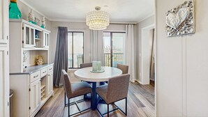 A dining area with a round table, chairs, a chandelier, a cabinet with decorative items, and a view through the window. - Beachfront condo w pool, tennis and pickleball, gym & balcony (Hilton Head)