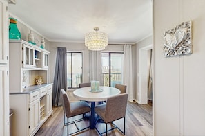 A dining area with a round table, chairs, a chandelier, a cabinet with decorative items, and a view through the window. - Beachfront condo w pool, tennis and pickleball, gym & balcony (Hilton Head)