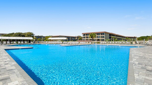 A large outdoor swimming pool with clear blue water, surrounded by a tiled deck and a building with multiple balconies in the background. - Beachfront condo w pool, tennis and pickleball, gym & balcony (Hilton Head)