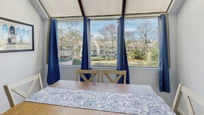 A room with a dining table, chairs, a patterned table runner, a framed photo on the wall, and a view through large windows with blue curtains. - Lagoon views, shared pool and easy access to Shelter Cove Towne Center (Hilton Head)