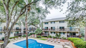 Un área de piscina con sillas de descanso y una alberca rodeada de árboles y un edificio de varios pisos con balcones.