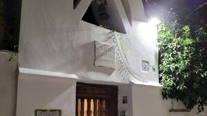 Un bâtiment blanc avec une porte en bois, une enseigne indiquant « ADE PARVULA DOMVS » et une petite fenêtre sur laquelle est inscrit le chiffre « 2 ».