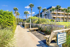 A wooden boardwalk leading to a building with balconies, surrounded by palm trees and greenery. - Newly Renovated Coastal Condo with Ocean-View Balcony, Shared Pool & Easy Beach Access (Hilton Head)