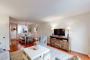 A living room with a white coffee table, a TV on a wooden stand, a dining area with a table and chairs, and a chandelier. - Updated Villa - Close to Coligny Plaza and Beach Access (Hilton Head)
