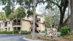 A multi-story building with balconies, a sign indicating '21 SANDS VILLAGE,' and a well-maintained landscaped area with trees and shrubs.