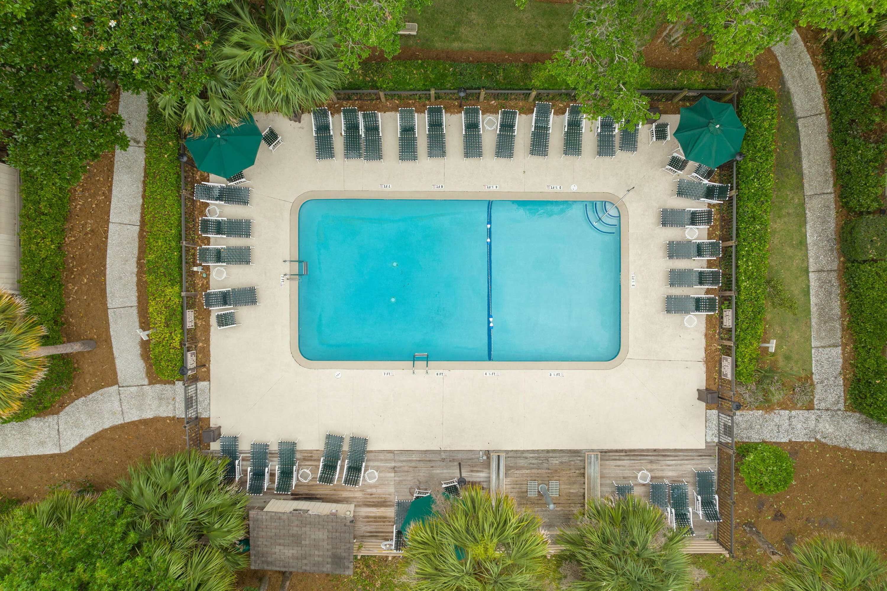Vue aérienne d’une piscine rectangulaire entourée de chaises longues et de parasols, avec une terrasse en bois située au fond.
