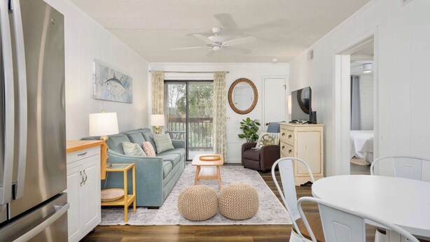 A modern living room with a sofa, coffee table, and ottomans, a dining area with a table and chairs, and a kitchen with a refrigerator and cabinets.
