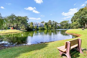 A serene lakeside view with a bench, a well-maintained lawn, and a row of houses in the background. - Cozy condo with balcony, pools, shared tennis and internet (Hilton Head)