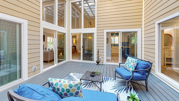 Eine überdachte Terrasse mit Holzboden, ein blaues Sofa mit gemusterten Kissen, ein kleiner Tisch mit einer Pflanze und Blick ins Haus durch Glastüren.