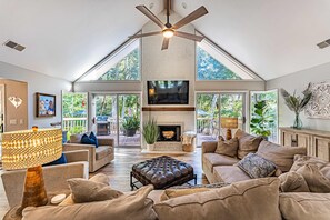A spacious living room with a fireplace, large windows, and a ceiling fan. - Palmetto Dunes house with private pool, shared tennis courts plus near beach & golf (Hilton Head)