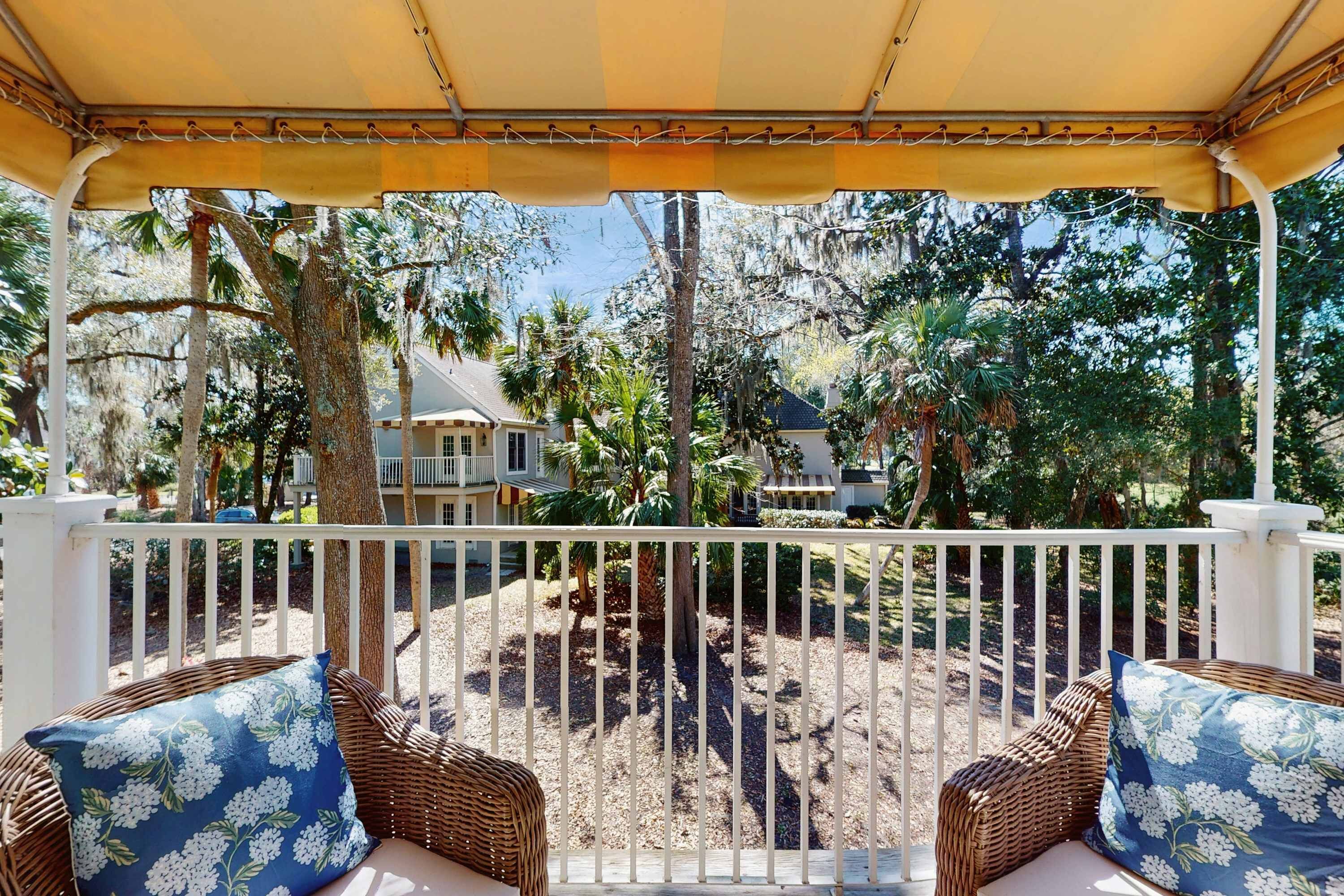 Uma área de estar coberta ao ar livre, com móveis de vime e guarda-corpo branco, com vista para um jardim ajardinado com árvores e uma casa.