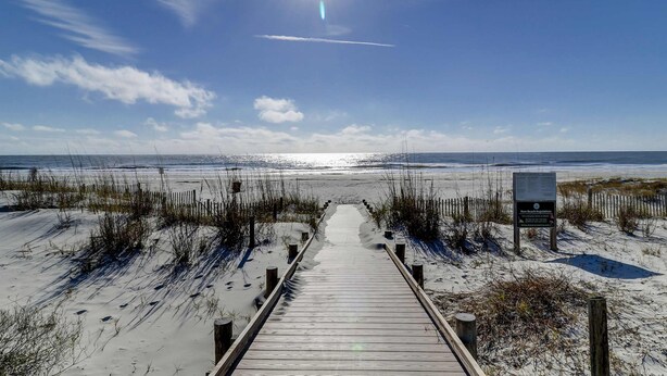 Ein hölzerner Steg führt zu einem Strand mit einem sandigen Pfad, einem klaren Himmel und einem Schild.