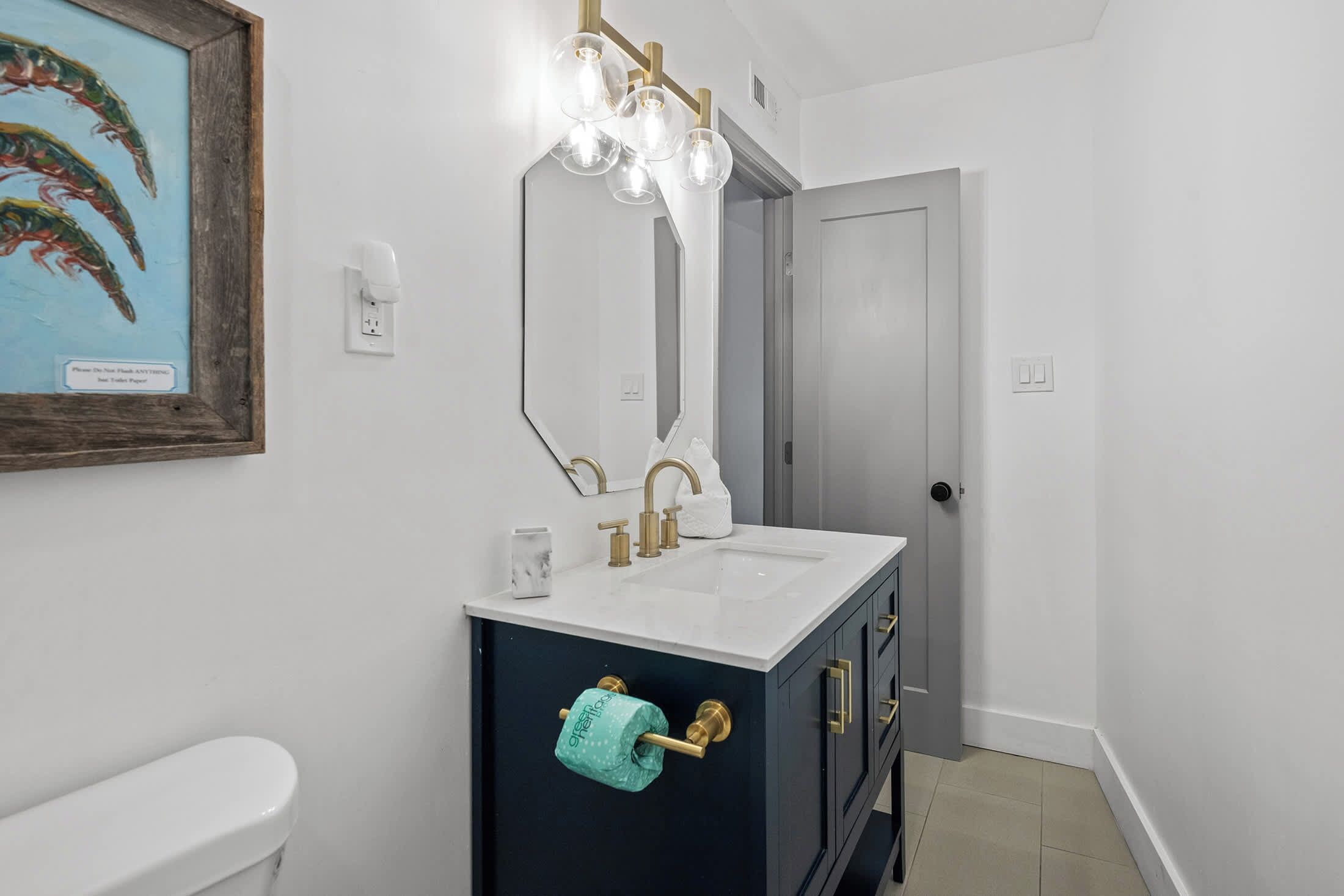 Ein Badezimmer mit einer weißen Toilette, einem dunklen Waschtisch mit Goldarmatur, einem Spiegel mit Beleuchtung und einer Tür.