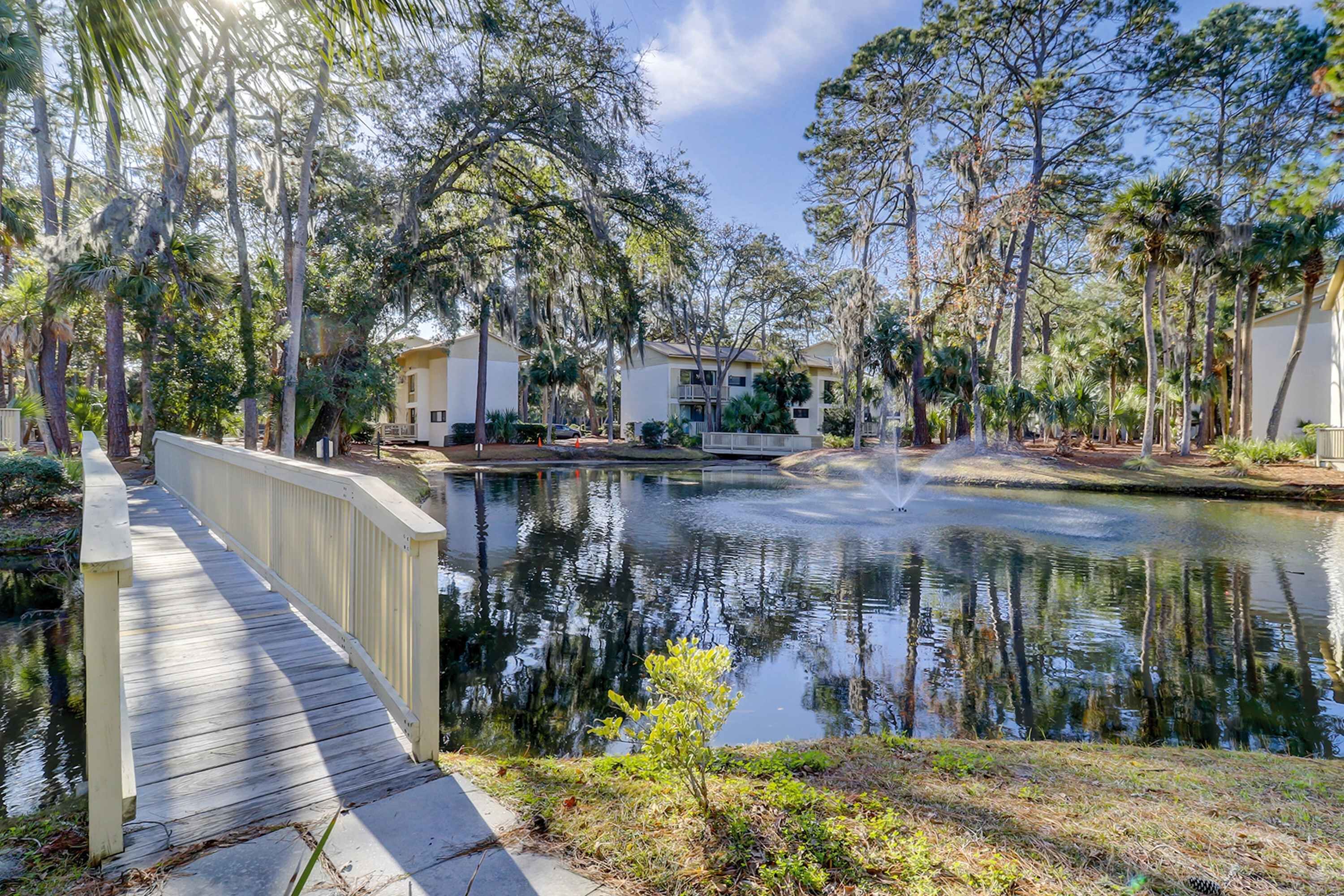 A wooden bridge over a calm water body with reflections of trees and buildings, surrounded by greenery and a clear sky.