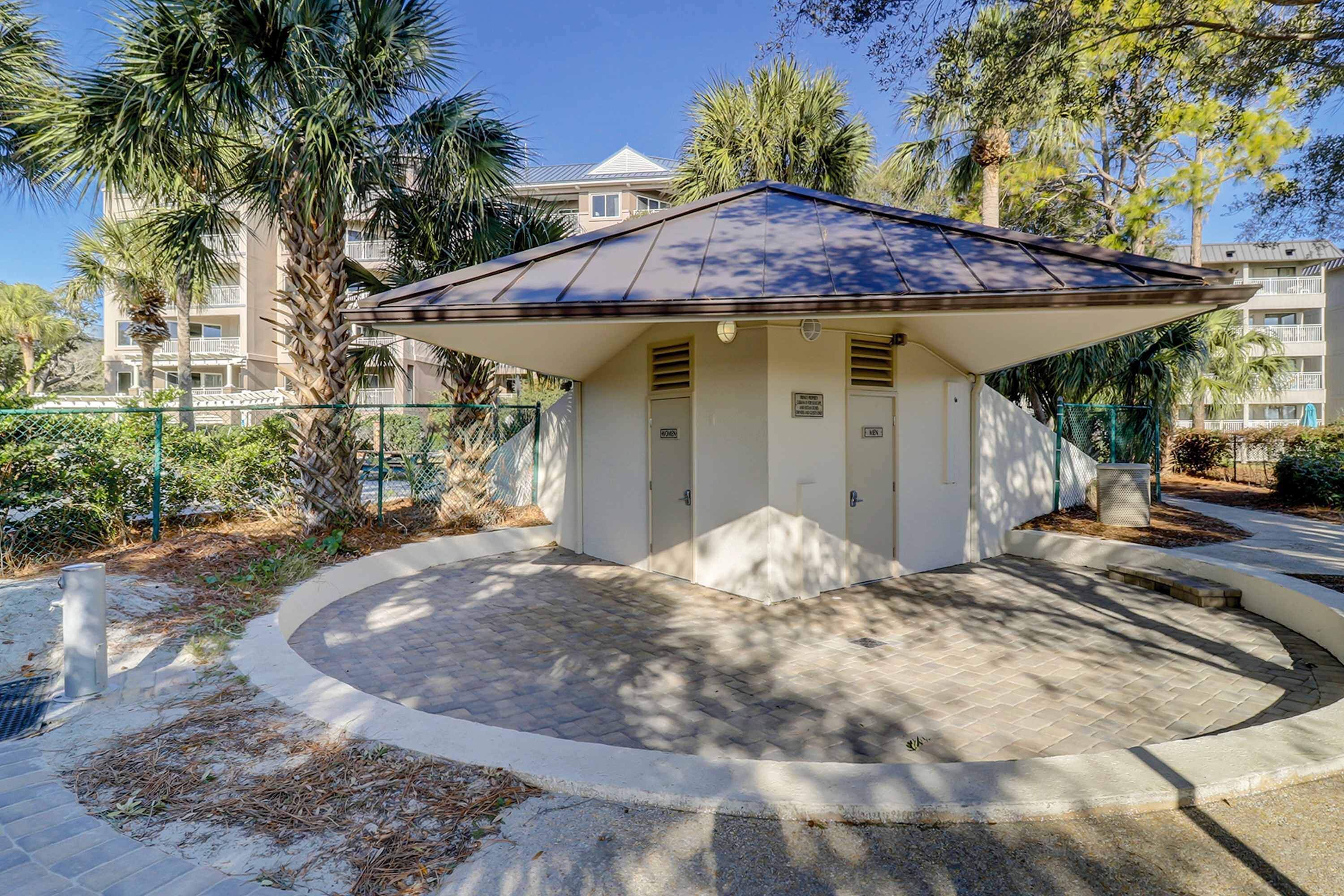 A small, single-story building with a sloped roof, surrounded by a paved area and palm trees.