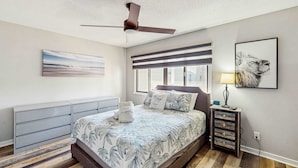 A bedroom with a bed, a ceiling fan, a window, a bedside table with a lamp, and a wall-mounted artwork.