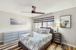 Ein Schlafzimmer mit einem Bett, einem Deckenventilator, einem Fenster, einem Nachttisch mit Lampe und einem an der Wand befestigten Kunstwerk.