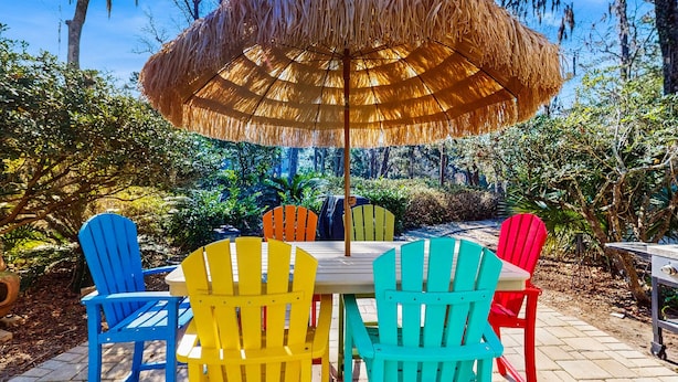 A patio with a table and colorful Adirondack chairs, a thatched umbrella, and a natural backdrop.